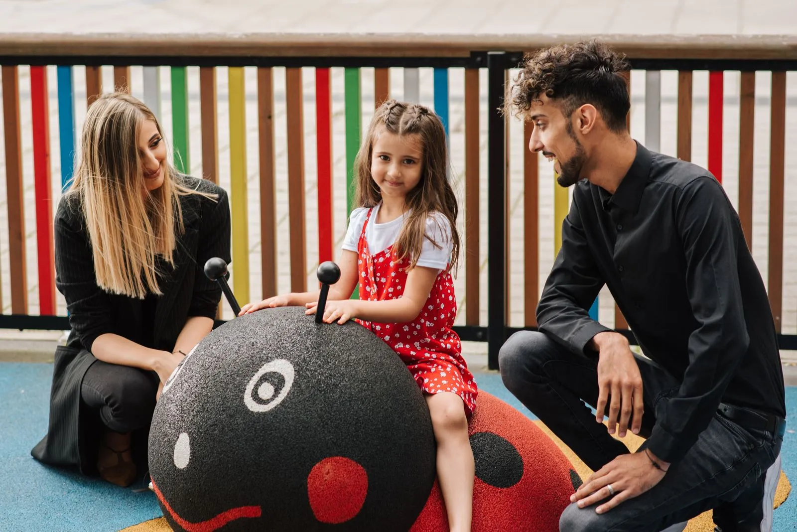 Little girl sitting on big toy ladybug on playground with mother and father sitting nearby and smiling